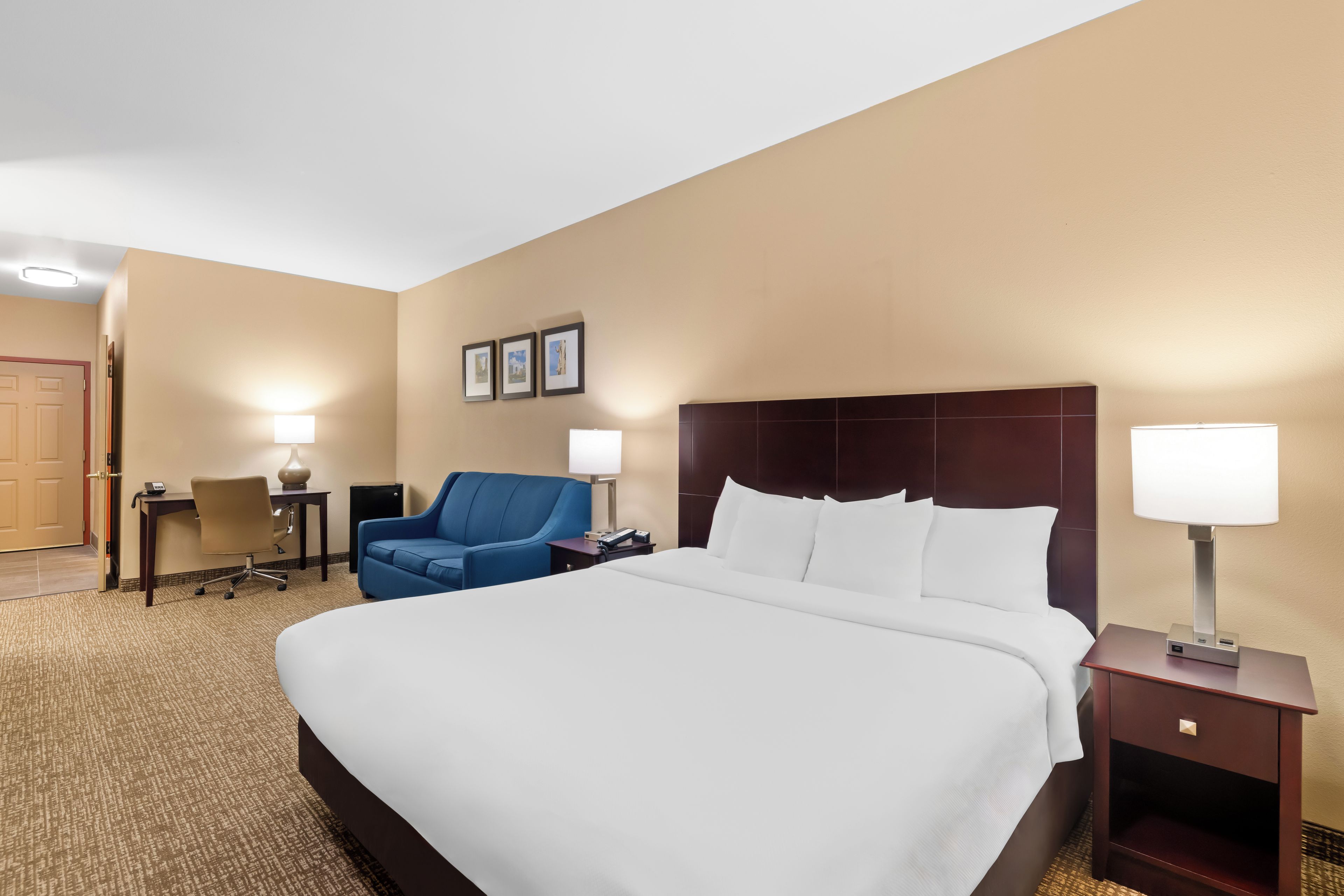 A hotel room with a large bed, a desk with a chair, a blue armchair, and two bedside lamps.