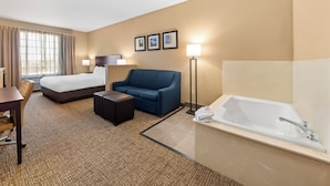 A hotel room with a bed, a sofa, a desk, a chair, a small table, a floor lamp, and a bathtub. - Roanoke Suites (Roanoke)