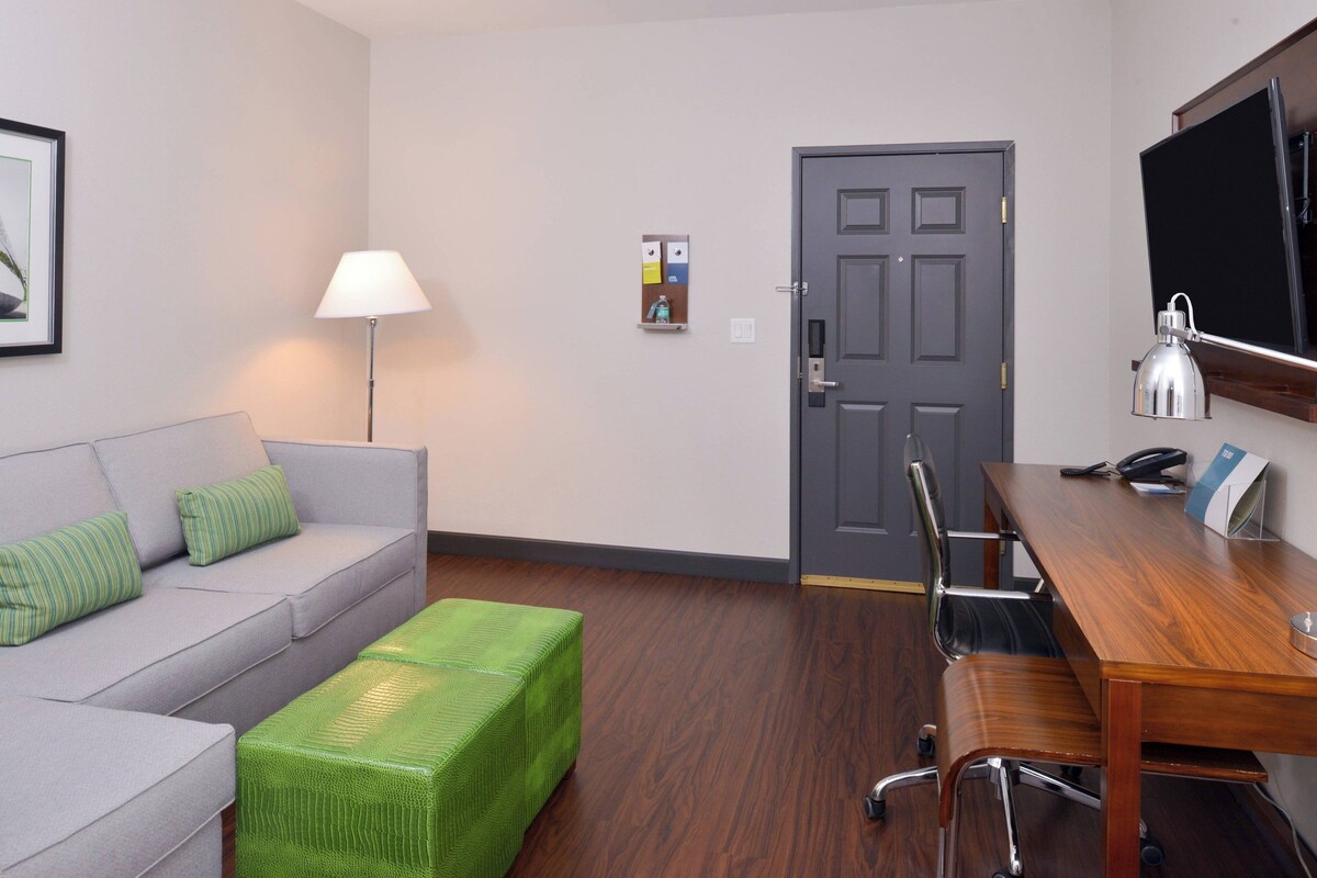 Una habitación con un sofá gris, cojines verdes, un otoman verde, un escritorio de madera con una lámpara y una puerta con un tablón de anuncios.