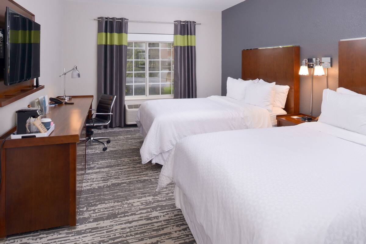 Quarto de hotel com duas camas, uma escrivaninha com cadeira, televisão e janela com cortinas.