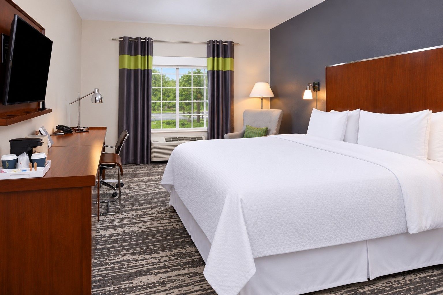 Habitación de hotel con una cama grande, un escritorio con teléfono y tazas de café, una silla, un televisor y una ventana con cortinas.