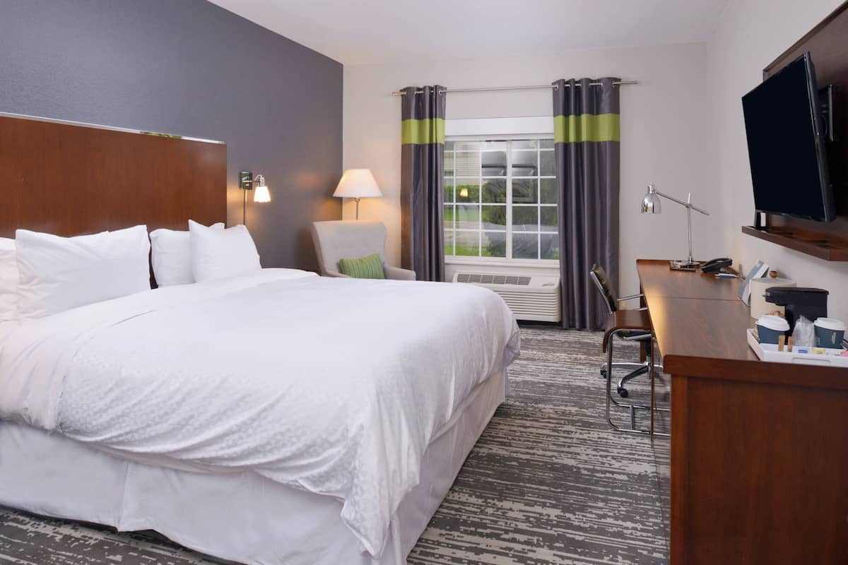 Quarto de hotel com uma cama grande, uma escrivaninha com um computador, uma cadeira, um abajur e uma televisão.