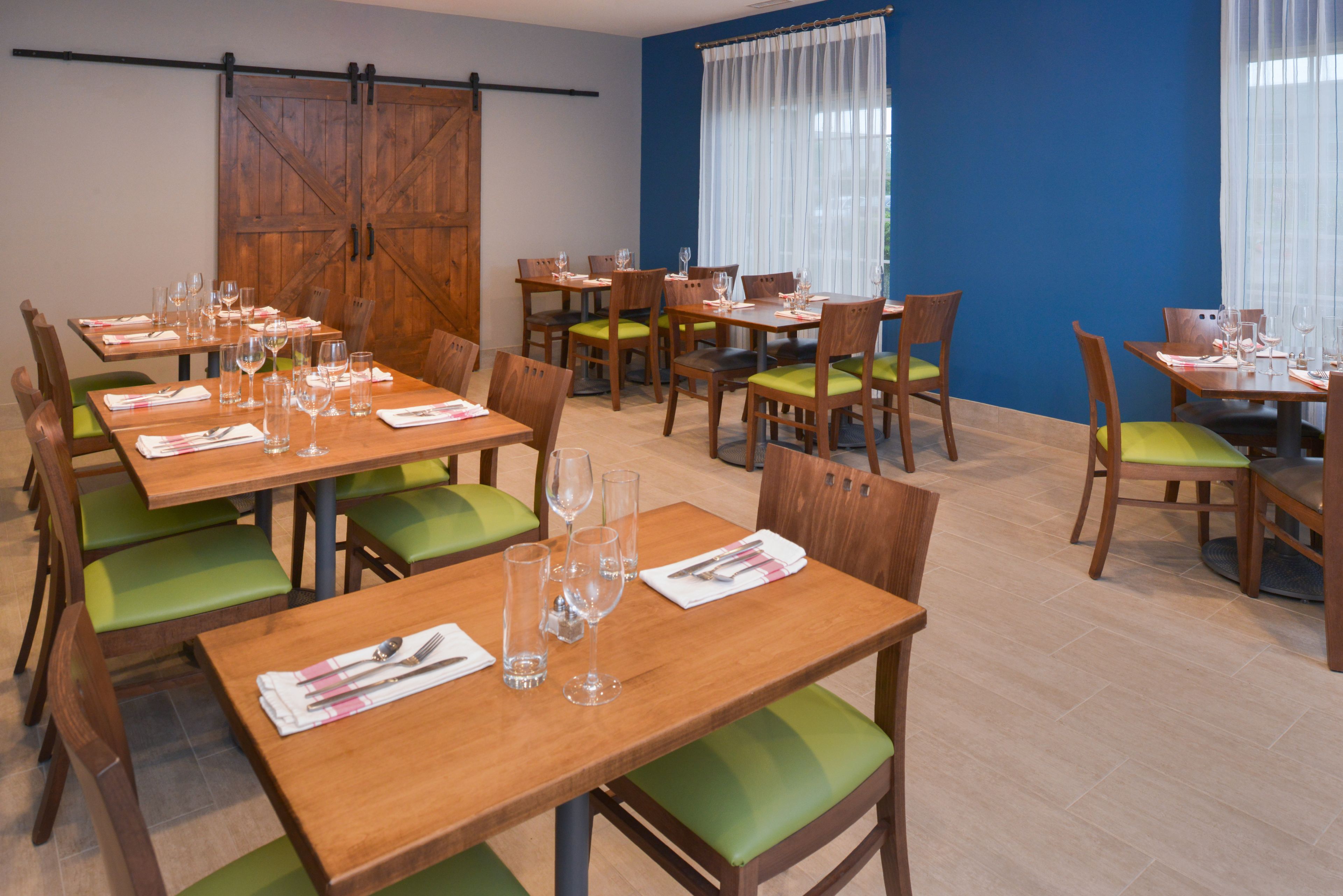 A dining area with multiple tables and chairs, wooden tables with green cushioned chairs, and a large wooden door in the background.