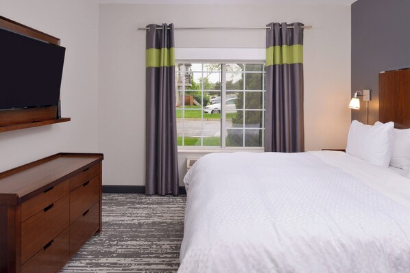 A hotel room with a bed, a wooden dresser, a flat-screen TV mounted on the wall, and a window with curtains.