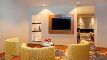 A hotel lobby with a flat-screen TV mounted on the wall, a small table with a decorative bowl, and three upholstered chairs.