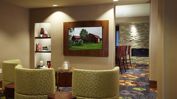 A hotel room with a large TV mounted on the wall, a wooden shelf with decorative items, and a carpeted floor. - Courtyard by Marriott Ithaca Airport/University (Ithaca)
