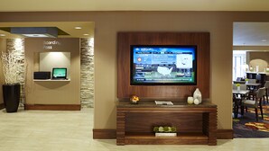 A hotel lobby with a television mounted on a wooden wall, a reception desk with a 'Boarding Pass' sign, and a seating area with tables and chairs. - Courtyard by Marriott Ithaca Airport/University (Ithaca)