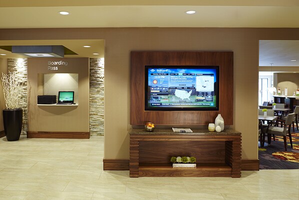 A hotel lobby with a television mounted on a wooden wall, a reception desk with a 'Boarding Pass' sign, and a seating area with tables and chairs.