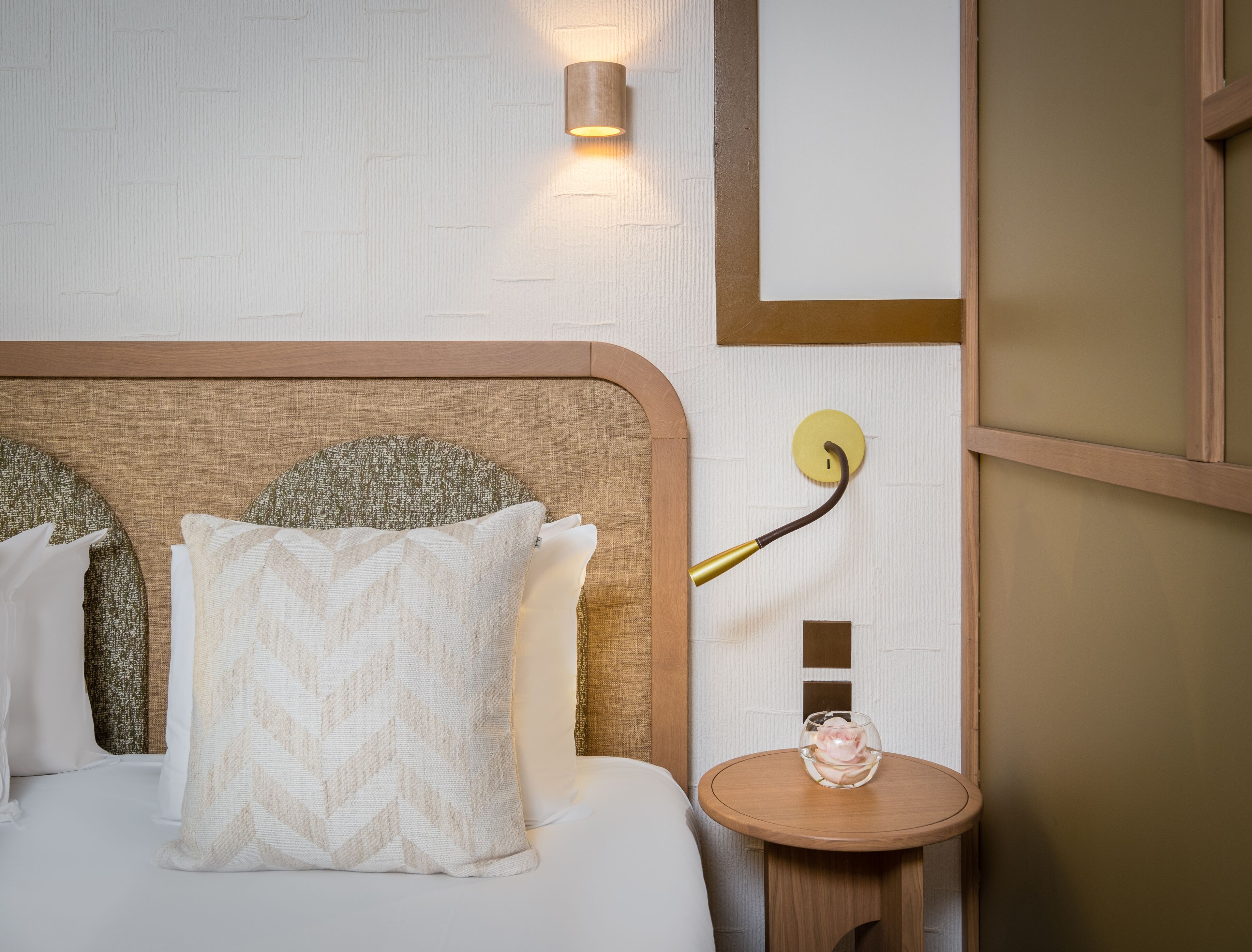 A neatly made bed with a headboard, a wall-mounted light fixture, and a small wooden table with a glass vase.