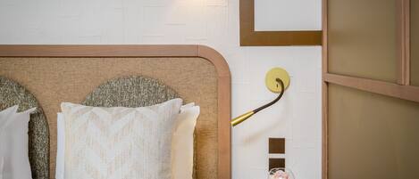 A neatly made bed with a headboard, a wall-mounted light fixture, and a small wooden table with a glass vase.