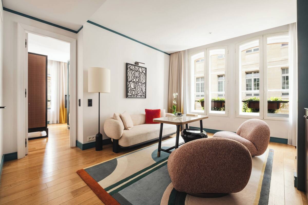 A modern hotel room with a sofa, two round chairs, a table, a floor lamp, and large windows.