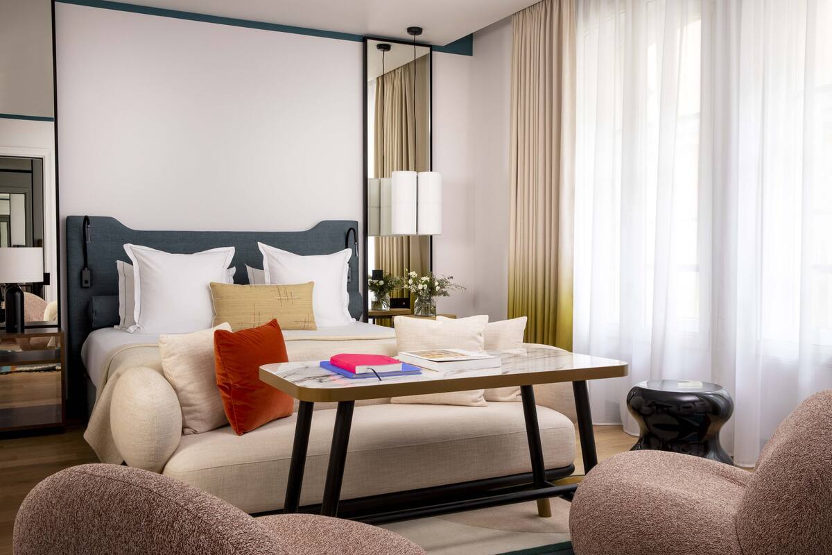 A hotel room with a bed, a bedside table with books, a lamp, and a view of the outside through sheer curtains.