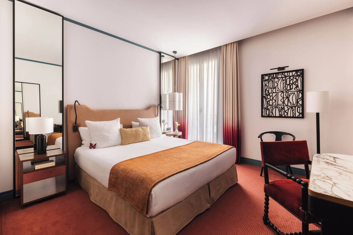A hotel room with a large bed, a bedside table, a chair, a desk, a lamp, and a mirror.