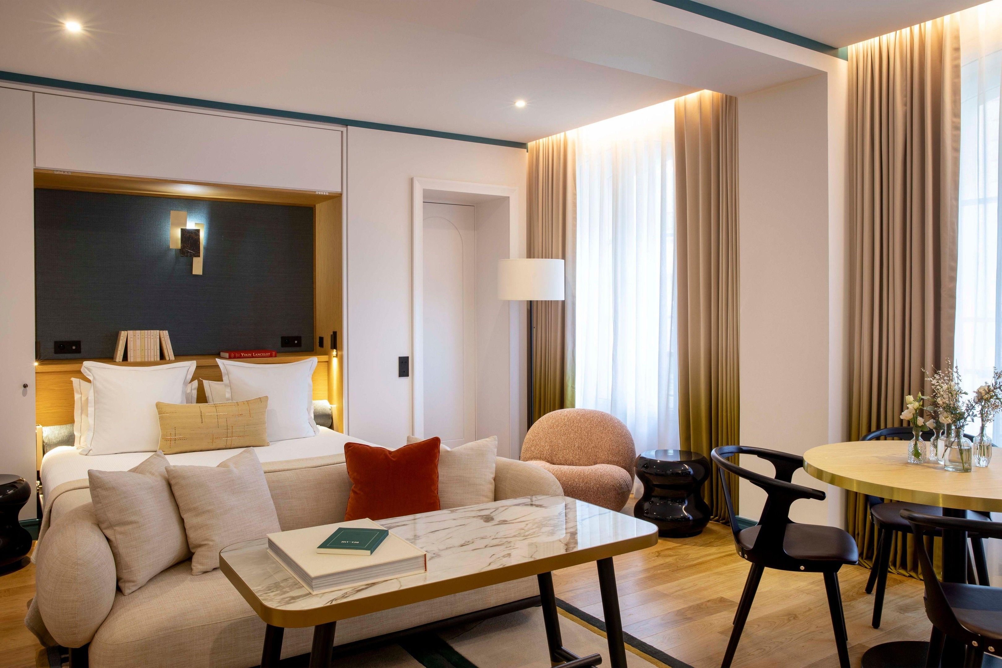 Una habitación de hotel moderna con sofá, una mesa de comedor con sillas, una lámpara y una estantería.