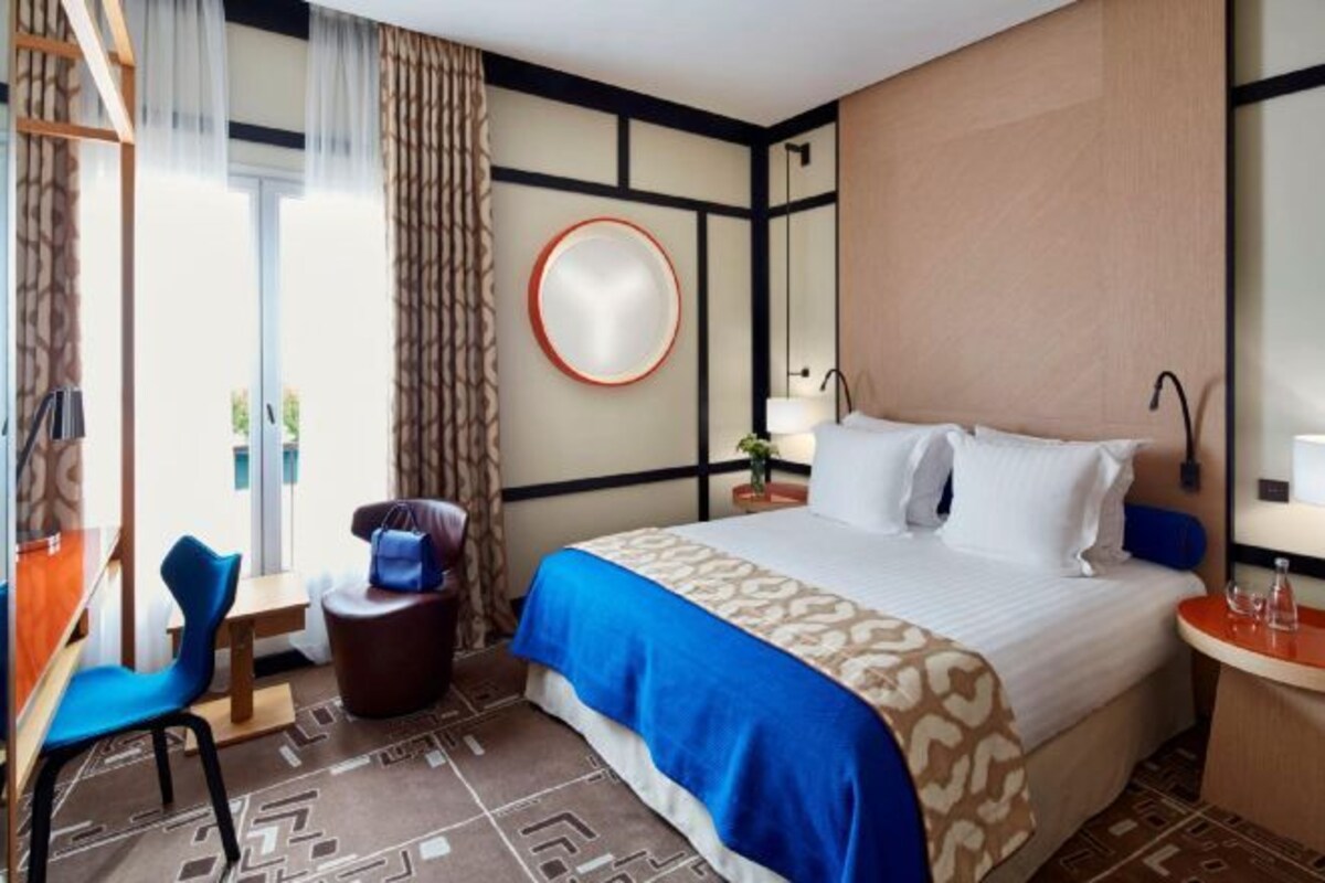 A hotel room with a bed, a desk, a chair, a round mirror, and a view of the outdoors.