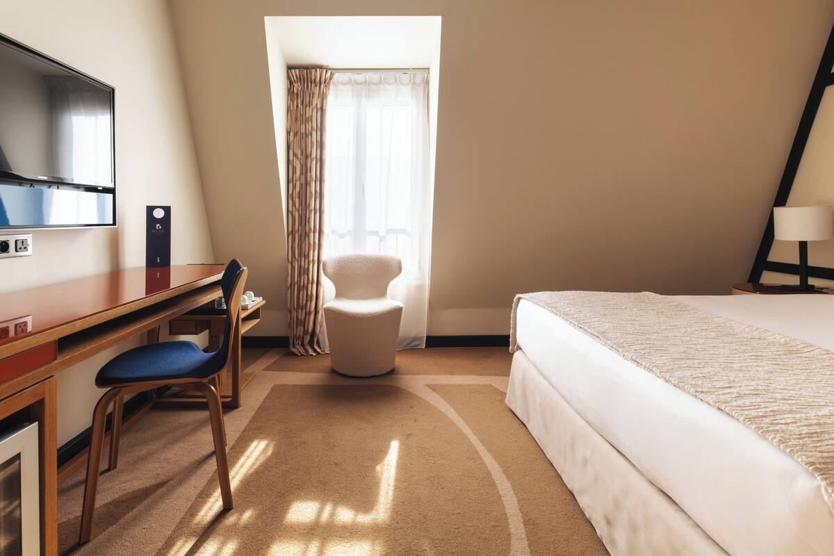 A hotel room with a bed, a desk with a chair, a television, a window with curtains, and a lamp.