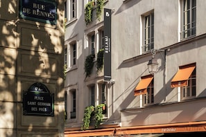 A building with a sign reading 'RUE DE RENNES' and another sign for 'BELAMI.' There are windows with orange awnings and some greenery on the facade.