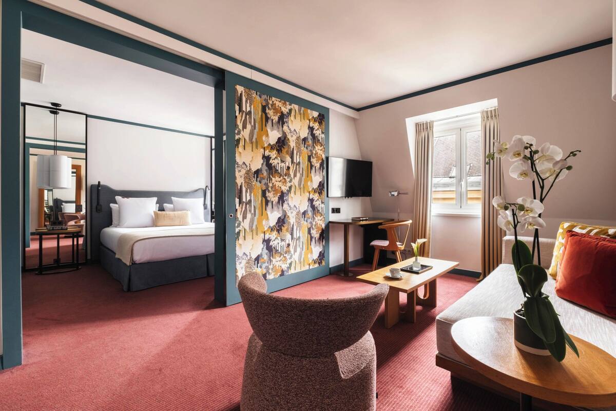 A hotel room with a bed, a desk, a chair, a TV, and a large abstract painting on the wall.