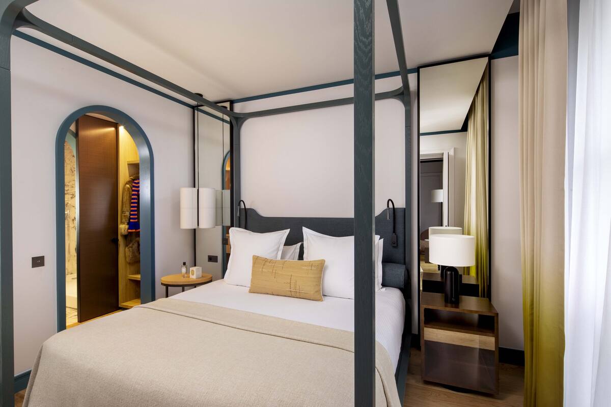 A modern hotel room with a four-poster bed, a bedside table with a lamp, and a wardrobe.