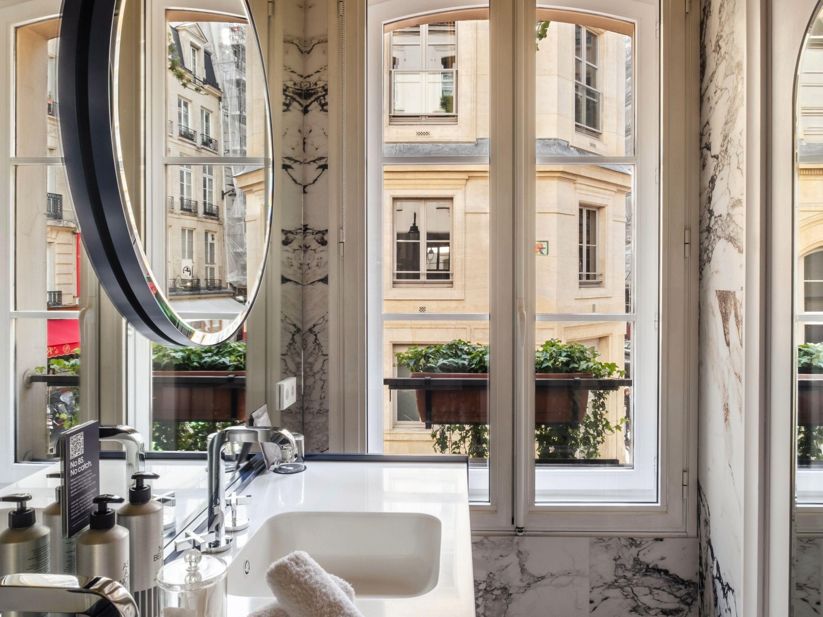 Une salle de bain dotée d’un grand miroir, d’un lavabo et offrant une vue sur un immeuble avec des balcons.