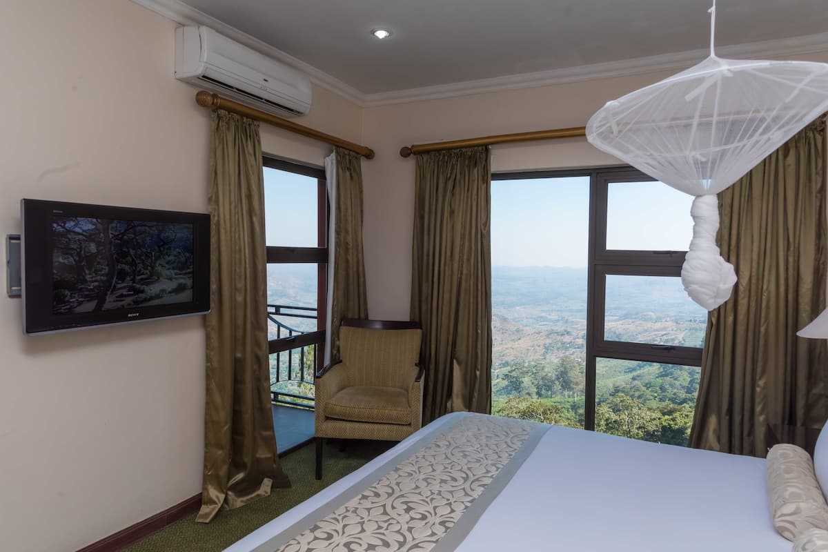 Quarto de hotel com cama, poltrona, televisão e janelas amplas com vista para a paisagem.