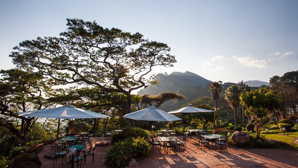 An outdoor dining area with tables and umbrellas, surrounded by lush greenery and a view of a mountain.