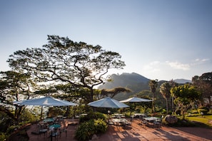 An outdoor dining area with tables and umbrellas, surrounded by lush greenery and a view of a mountain. - Sunbird Ku Chawe Hotel (Zomba)