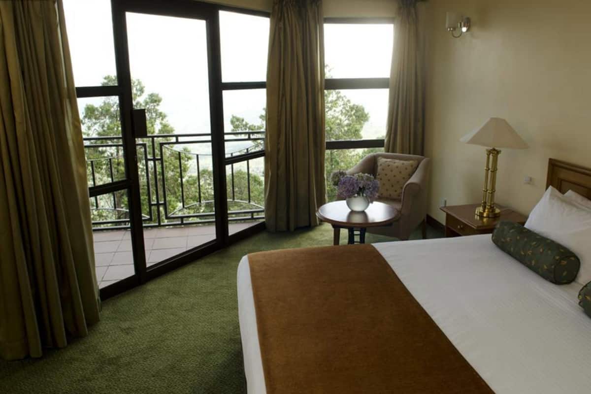 Quarto de hotel com uma cama, uma cadeira, uma mesinha com um vaso de flores e uma varanda com cortinas.