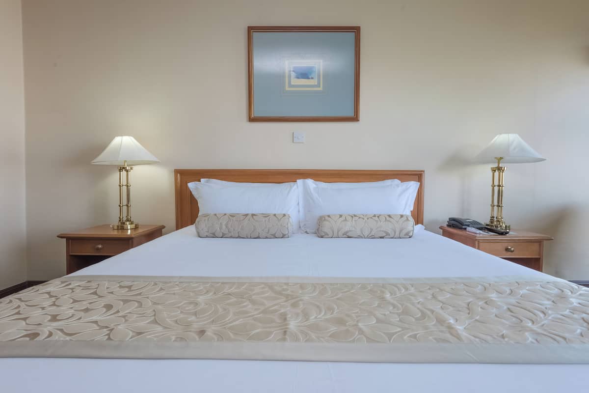 Uma cama bem arrumada com cabeceira decorativa, duas mesas de cabeceira com abajures e um quadro na parede.