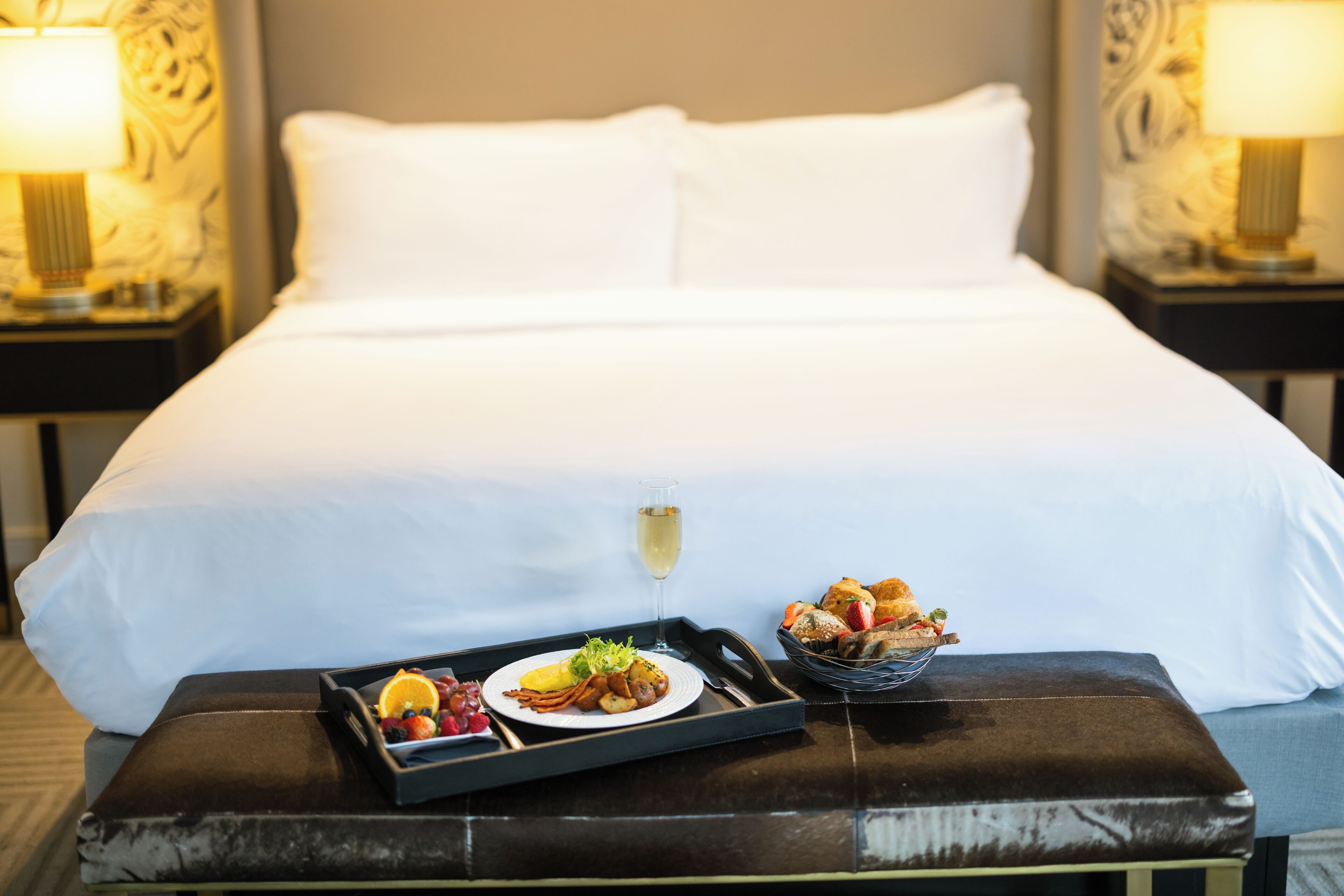 A hotel room with a neatly made bed, a tray with breakfast items, and a glass of champagne.