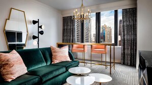 A modern hotel room with a green sofa, two round tables, a city view, and a chandelier.