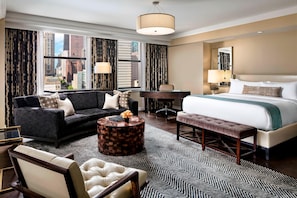 A hotel room with a large bed, a sofa, a desk, and a chair. There is a view of the city through the windows.