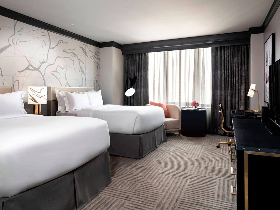 A hotel room with two beds, a desk, a chair, a lamp, and a large window with curtains.