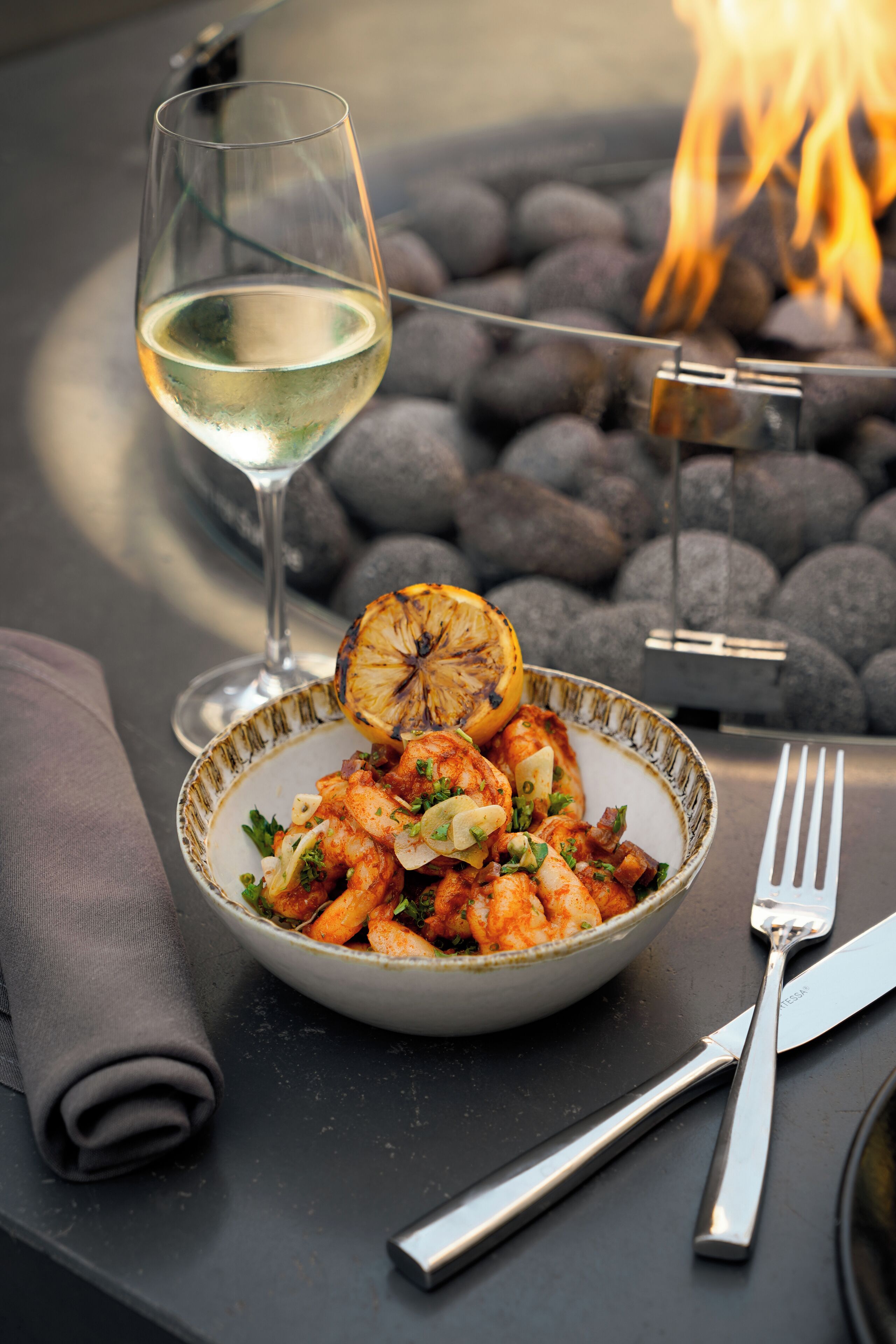 A bowl of food with a glass of white wine and a fork on a table with a fire in the background.