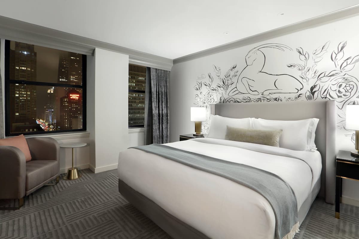 Habitación de hotel con una cama grande, una mesita de noche, una silla, un ventanal con vista a la ciudad y un mural decorativo.