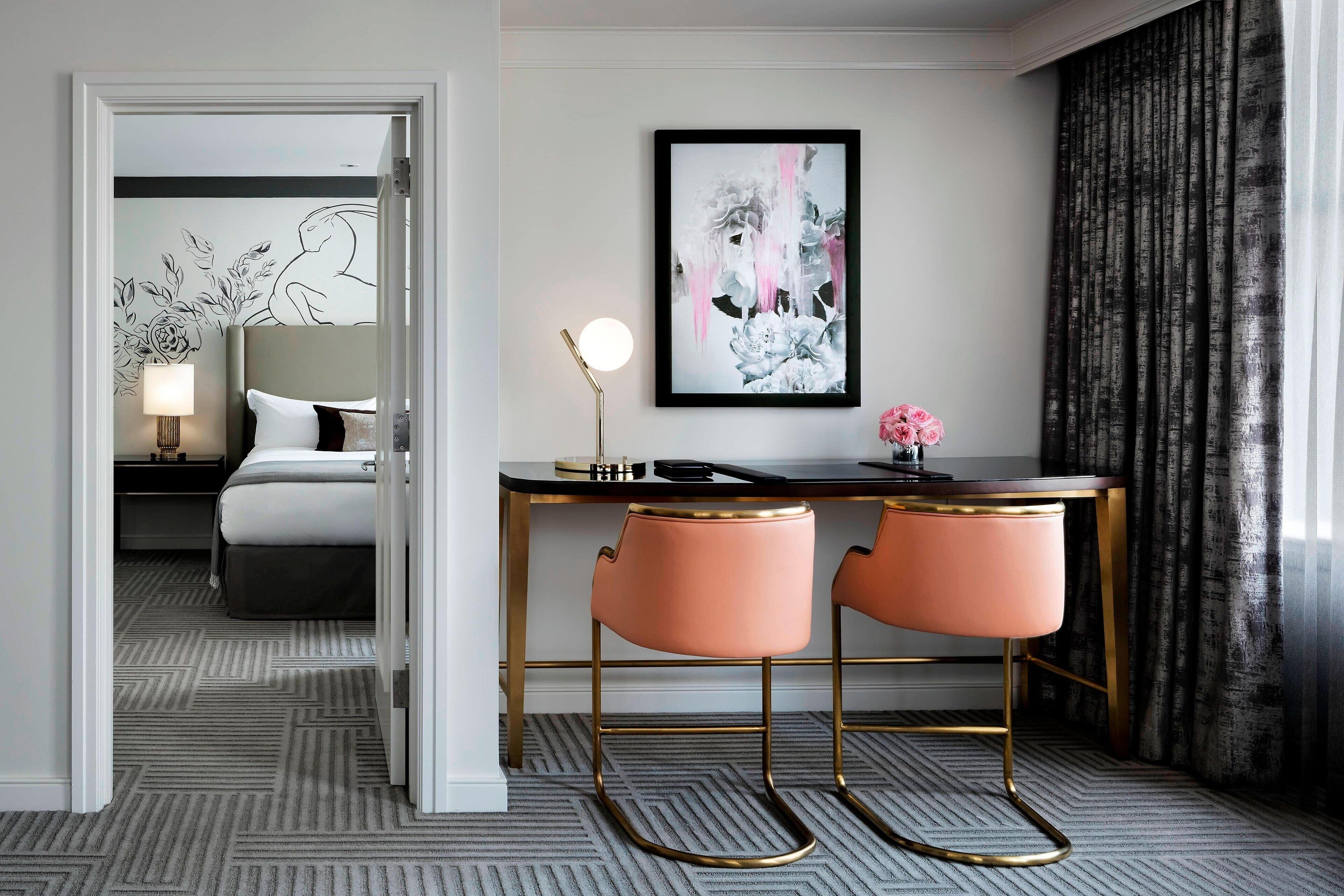 Una habitación de hotel con una cama, un escritorio con dos sillas rosas, una obra de arte enmarcada y una lámpara.