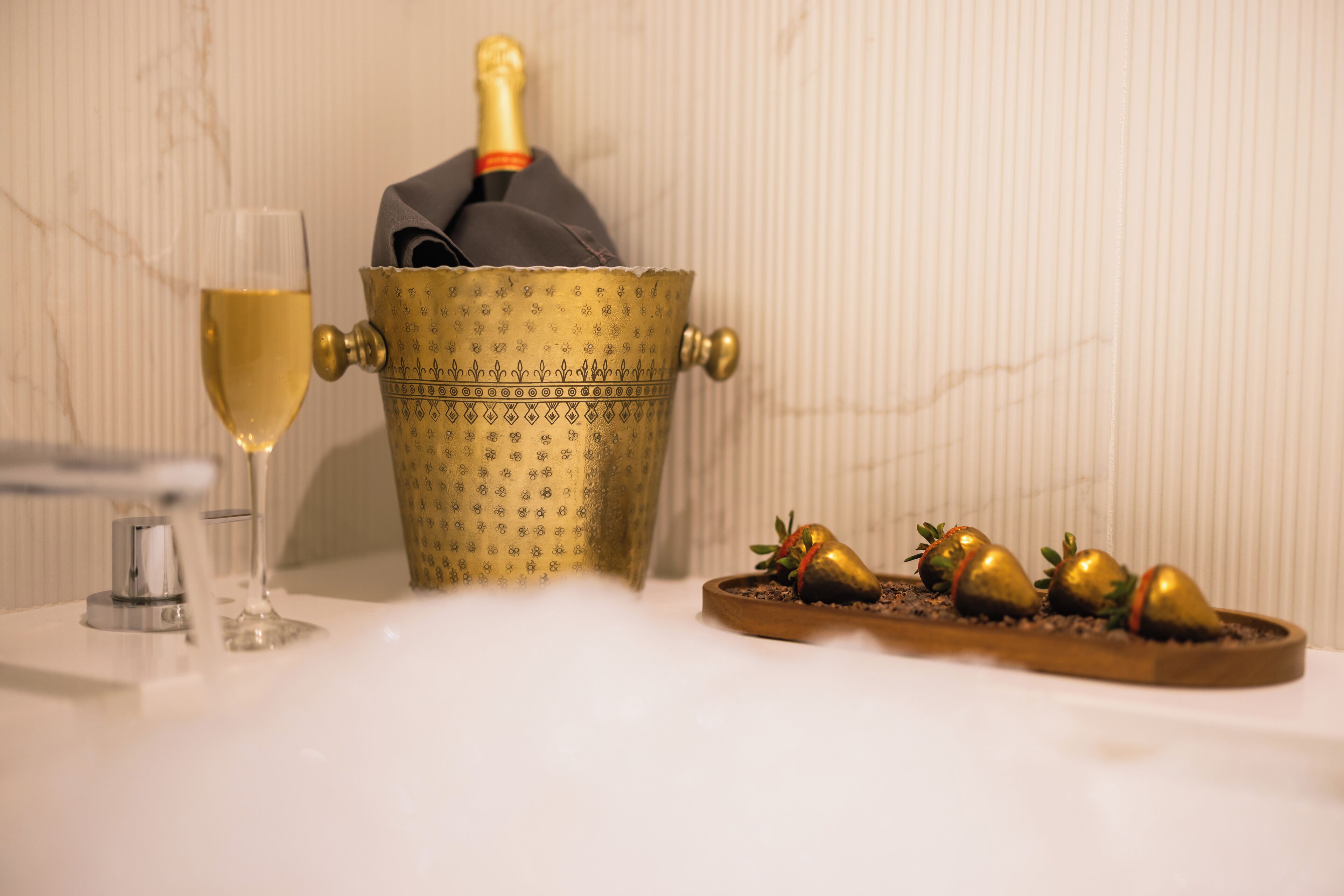 A champagne bottle in a decorative gold bucket, a glass of champagne, and a tray with golden strawberries.