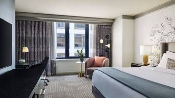 A hotel room with a bed, a desk, a chair, a small table with a lamp, and a view of the city through large windows.
