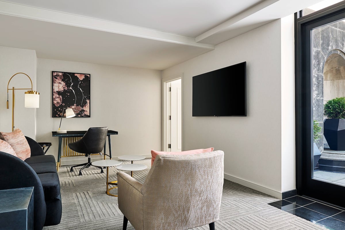 Una sala de estar moderna con un sofá, un sillón y un escritorio con una silla. Un televisor de pantalla plana está montado en la pared, y un ventanal ofrece vistas al exterior.