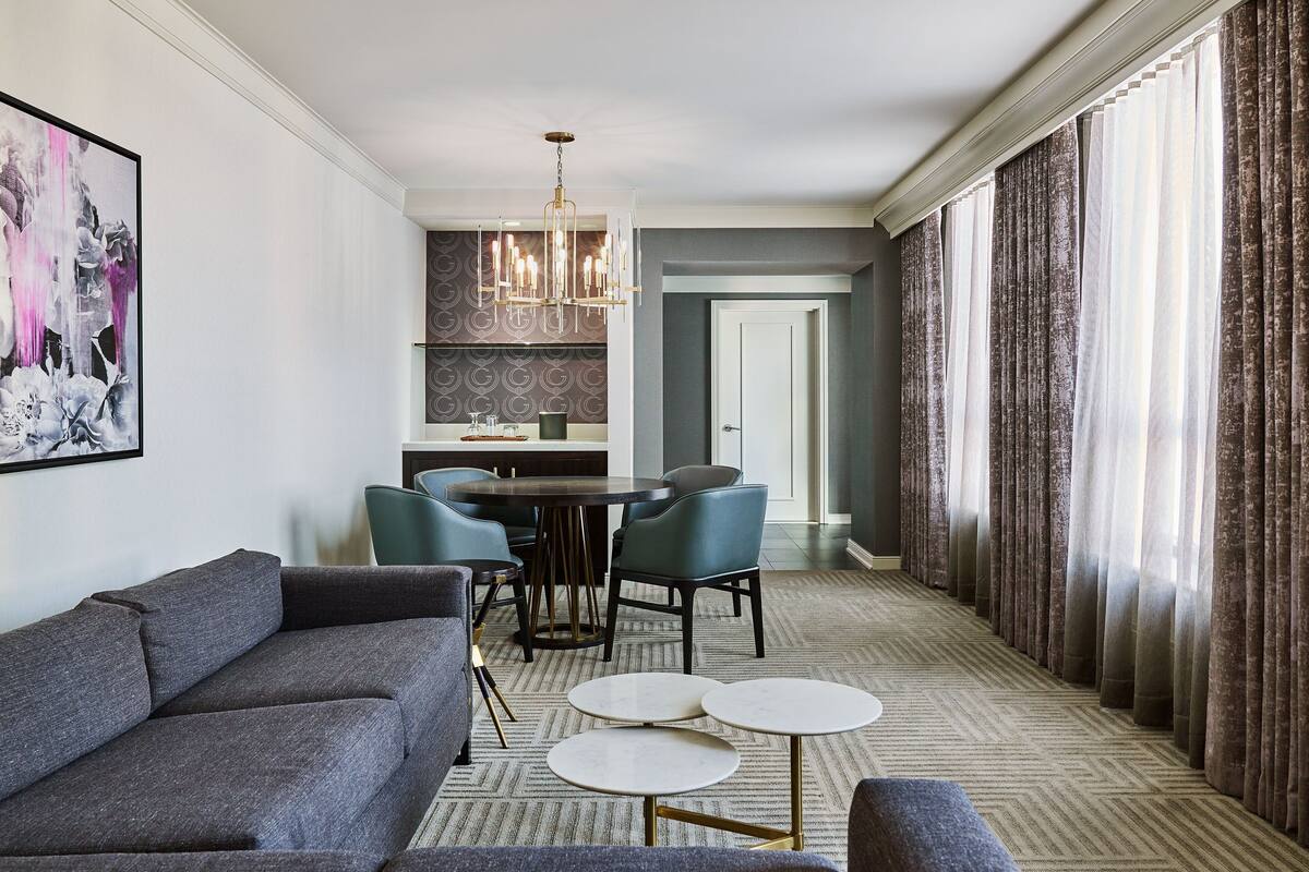 Una habitación de hotel moderna con un sofá gris, una mesa de comedor con sillas y una araña.