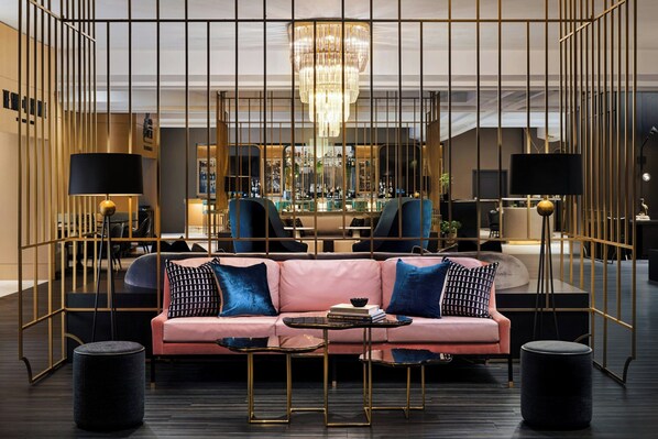 A modern hotel lobby with a pink sofa, black and blue pillows, a chandelier, and a bar area in the background.