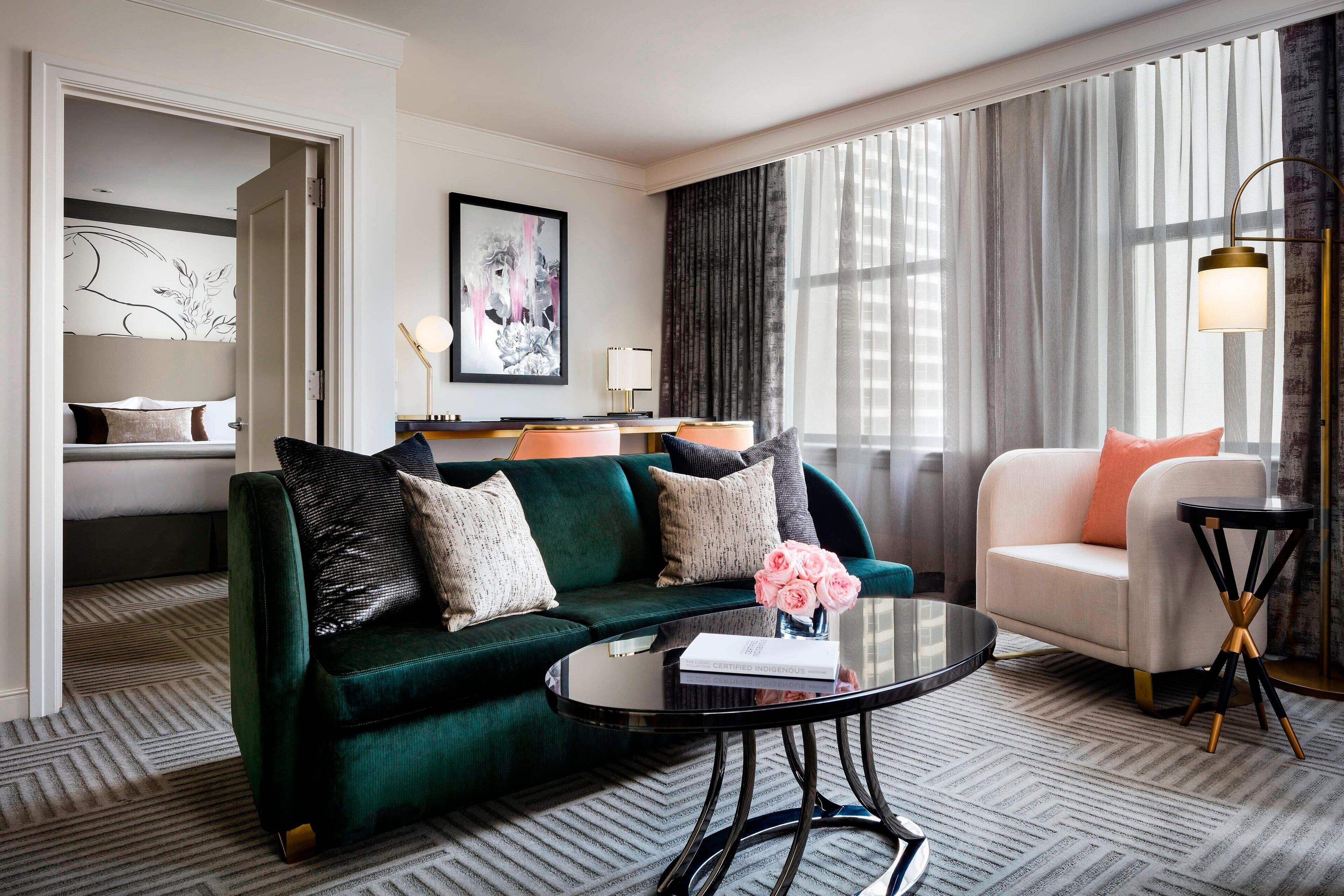 A modern hotel room with a green sofa, a white armchair, and a round coffee table. There is a painting on the wall and a window with curtains.