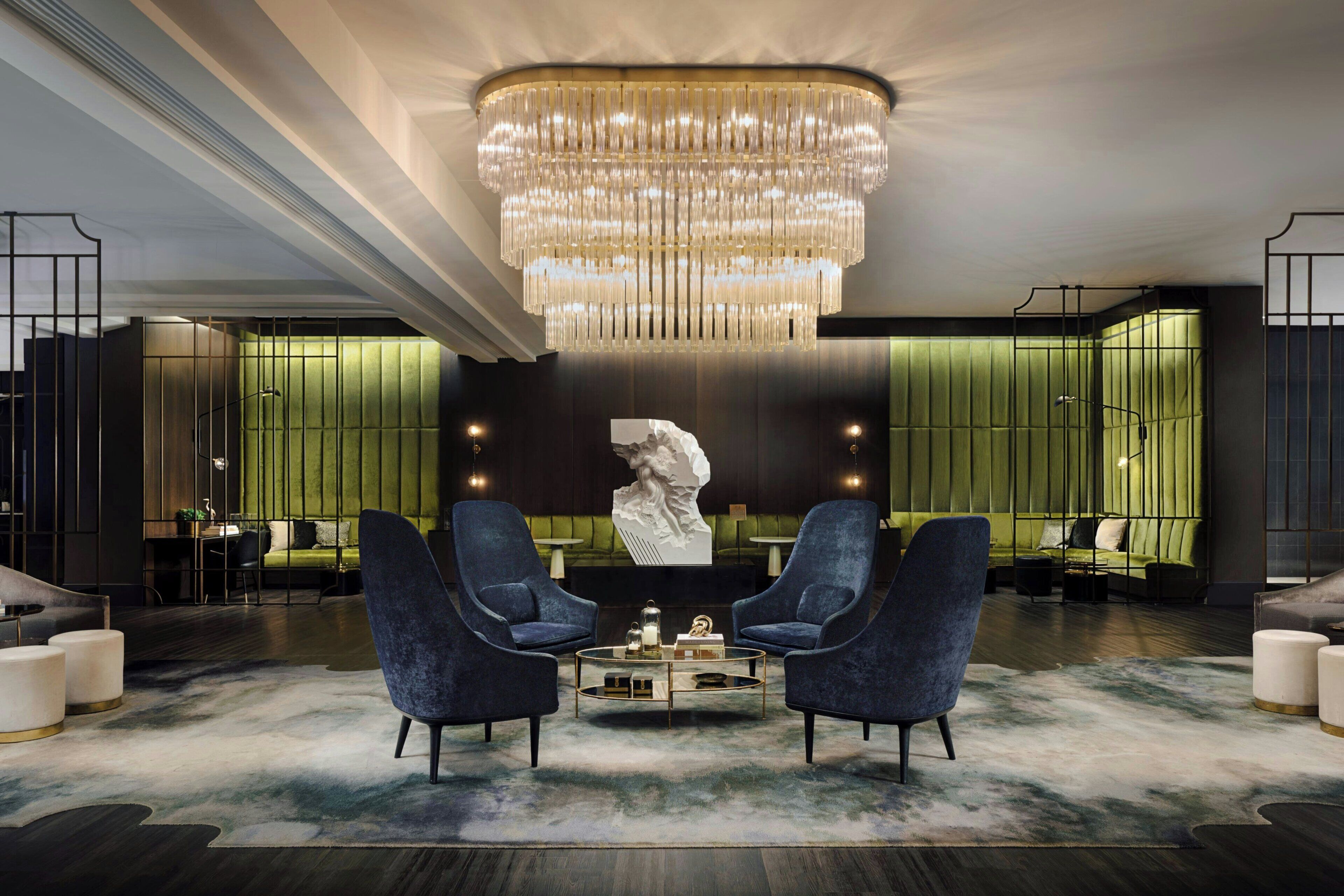 Um hall moderno com um grande candelabro, duas poltronas azuis, uma mesa de centro redonda e uma parede verde decorativa.