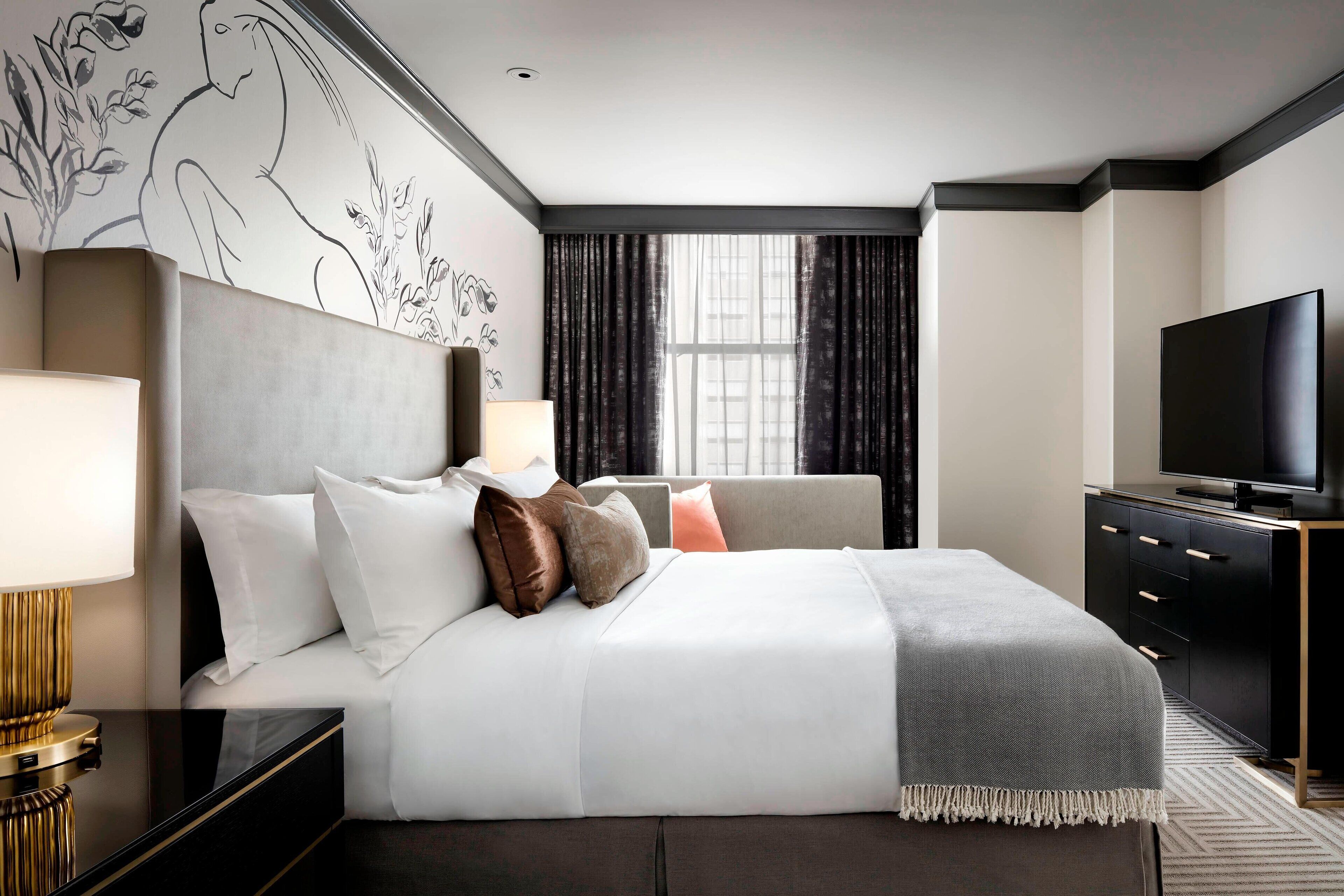 A modern hotel room with a large bed, a flat-screen TV, a desk with a lamp, and a large window with curtains.