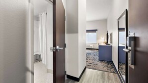 Um corredor de hotel com piso de madeira, um banheiro com cortina de chuveiro e uma mesa com luminária.
