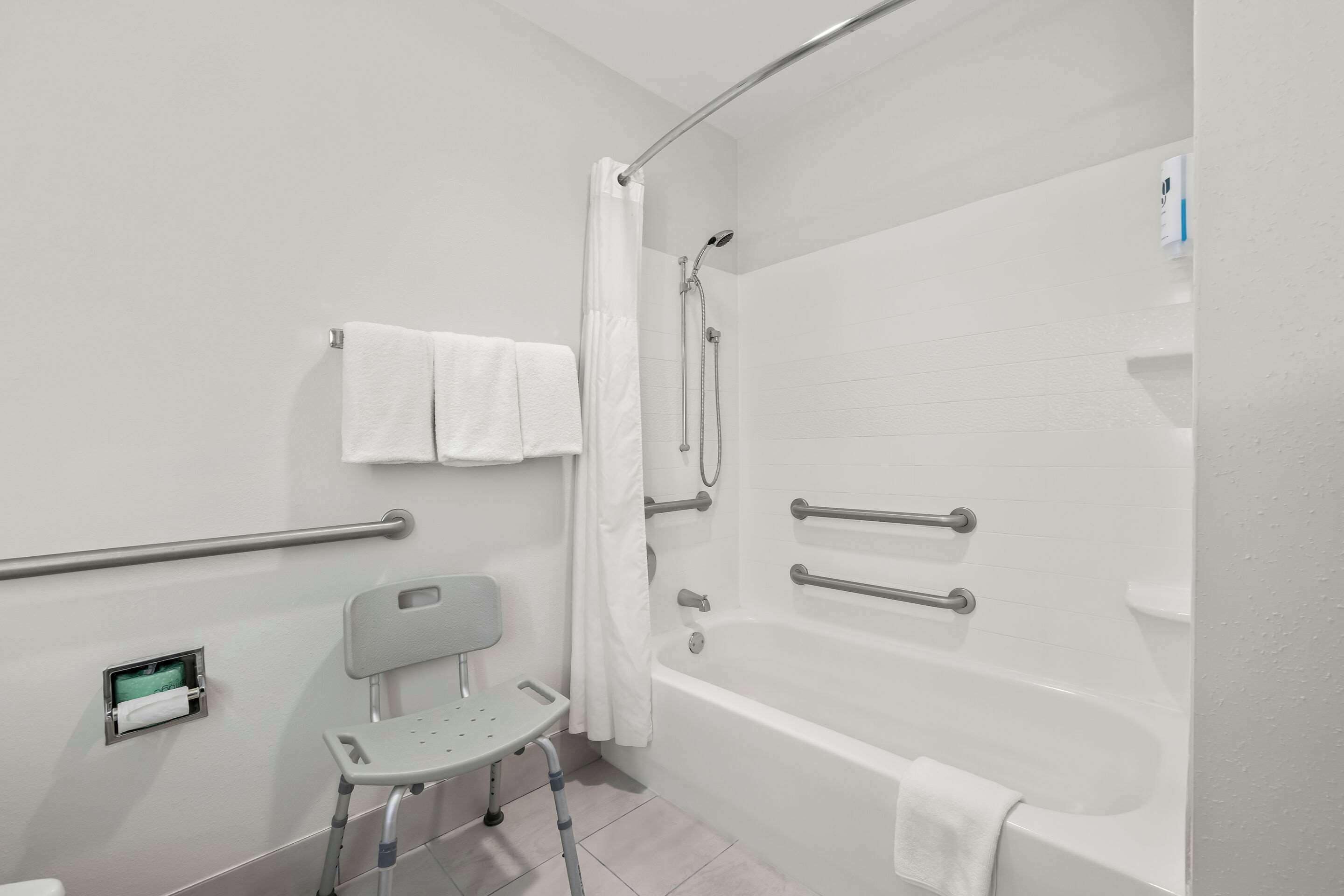 A bathroom with a bathtub, grab bars, a wheelchair-accessible chair, and a towel rack.
