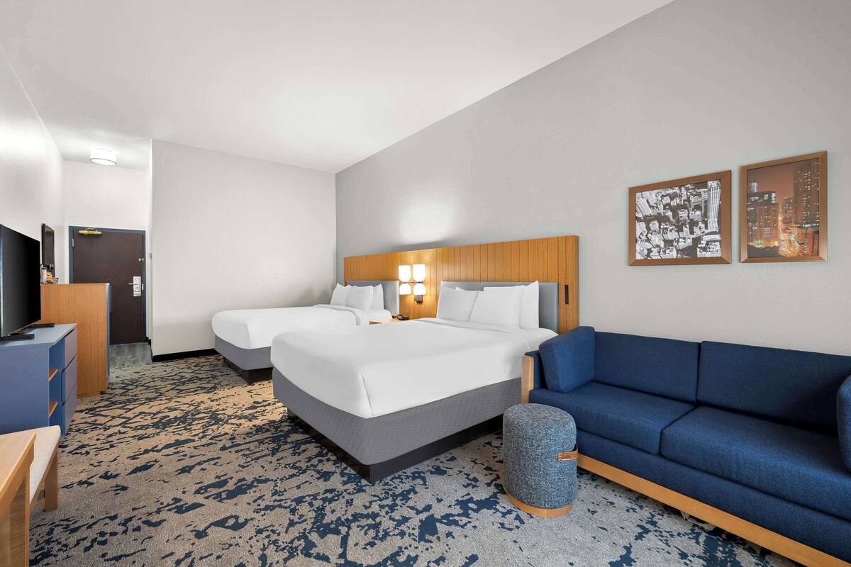 Habitación de hotel con dos camas, un sofá azul, un pequeño otoman y un escritorio con una silla.