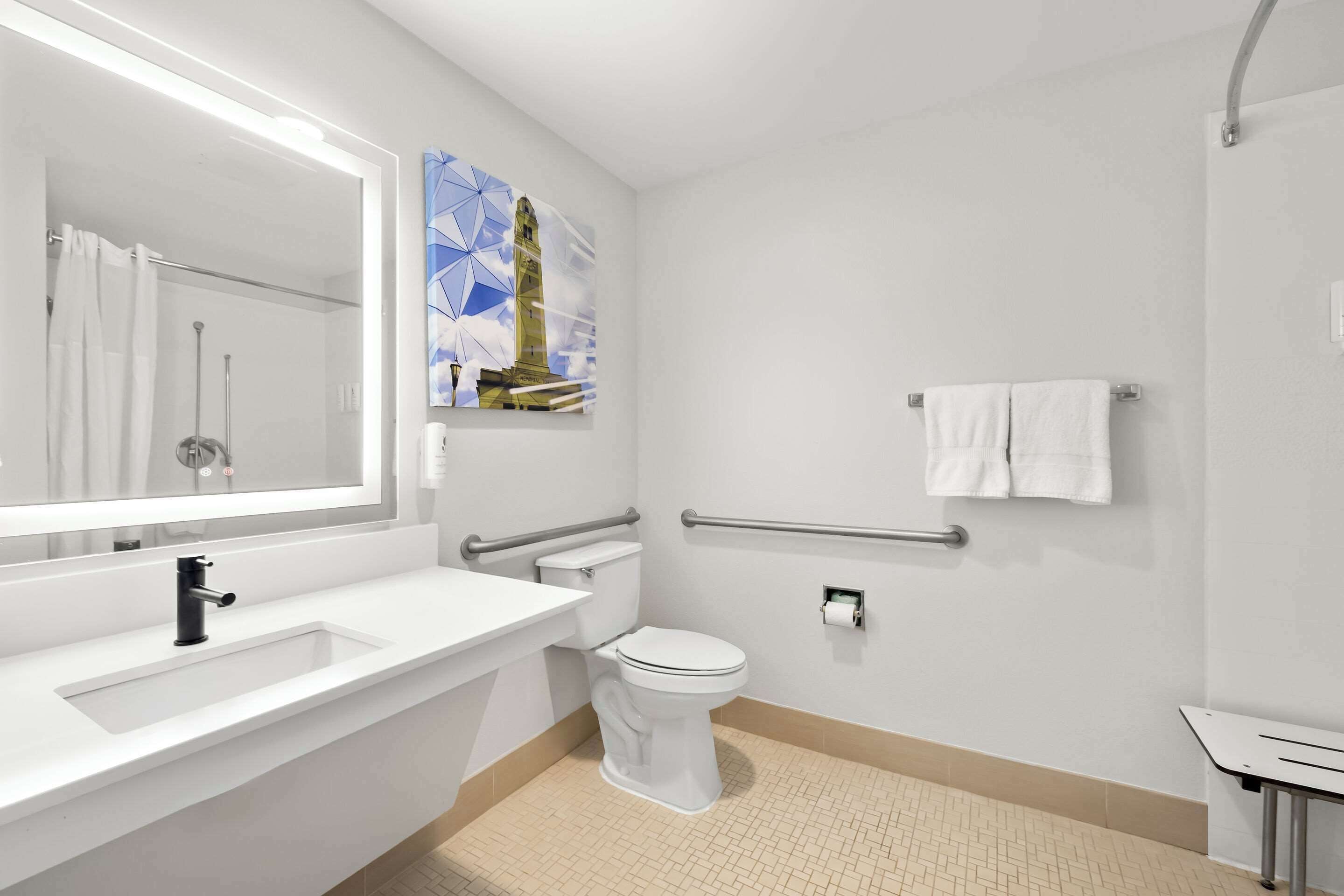A modern bathroom with a white sink, toilet, and mirror. There is a handrail and a towel rack on the wall.