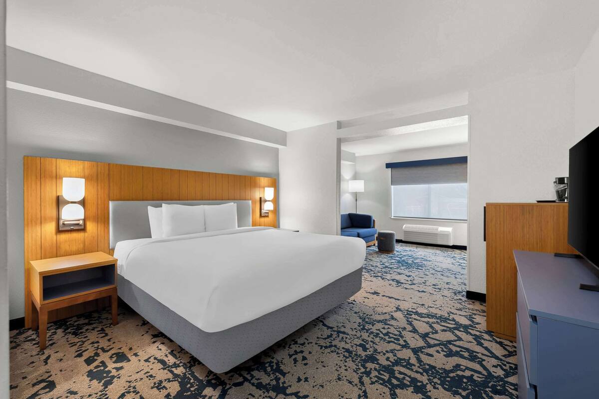 Una habitación de hotel con una cama grande, cabecera de madera, lámparas de noche, un escritorio con televisor y una alfombra azul estampada.
