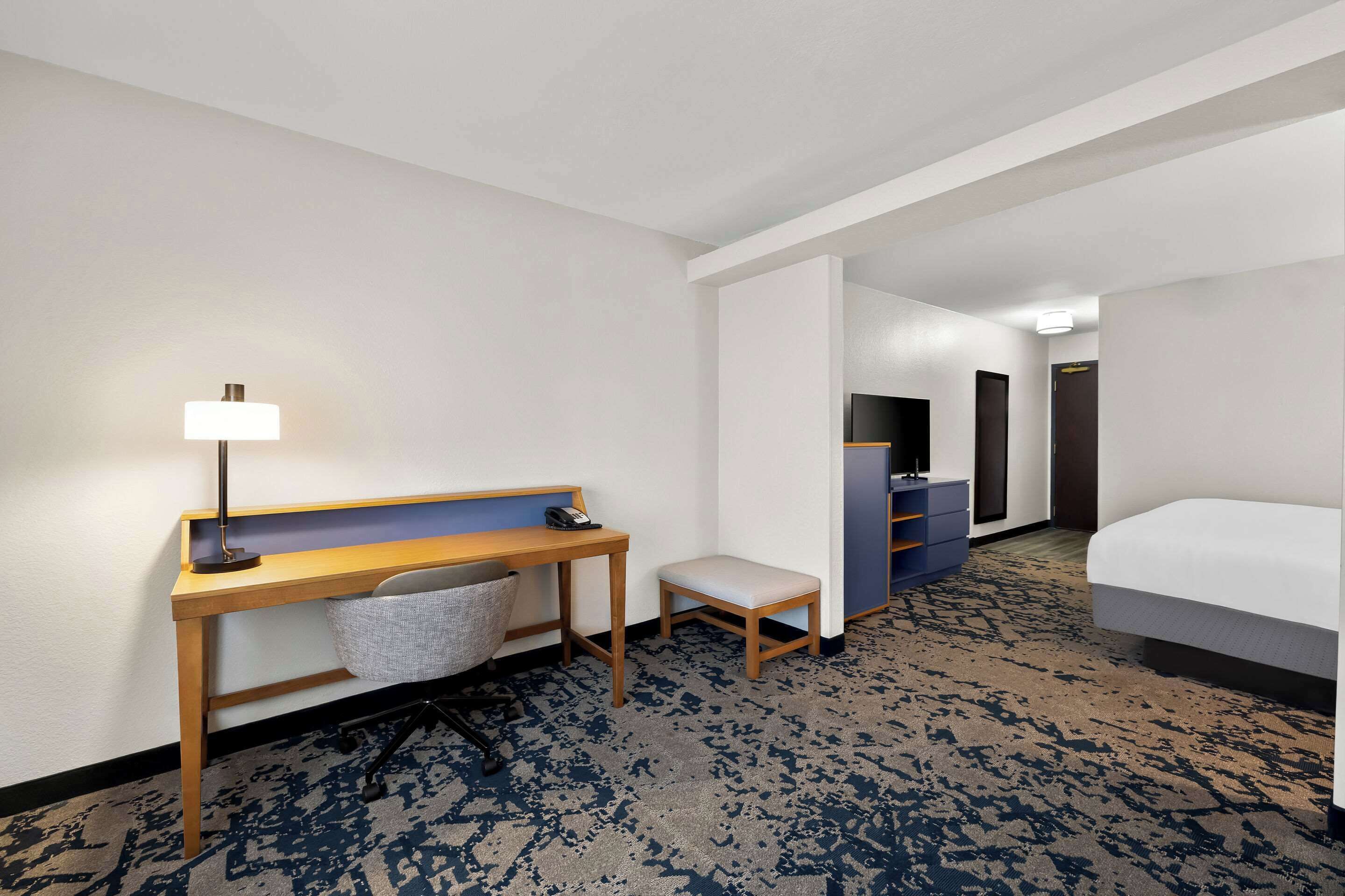 A hotel room with a bed, a desk with a lamp, a chair, a small bench, and a wardrobe.